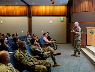 Rear Adm. William Greene, right, stands at the front of an auditorium speaking to a room of Sailors and civilians.