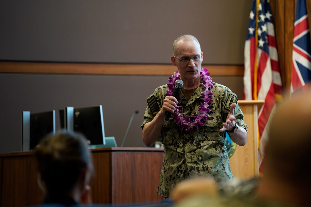Rear Adm. William Greene stands at the front of an auditorium speaking to a room of Sailors and civilians.