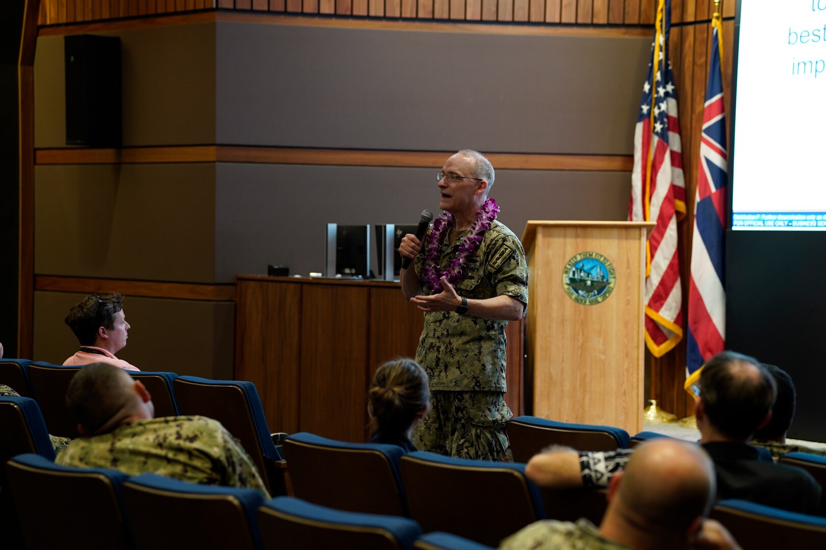 Rear Adm. William Greene stands at the front of an auditorium speaking to a room of Sailors and civilians.