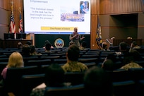Rear Adm. William Greene stands at the front of an auditorium speaking to a room of Sailors and civilians.