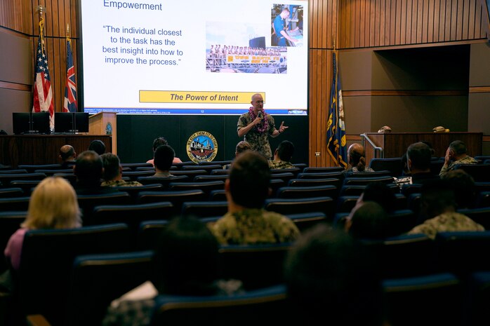 Rear Adm. William Greene stands at the front of an auditorium speaking to a room of Sailors and civilians.