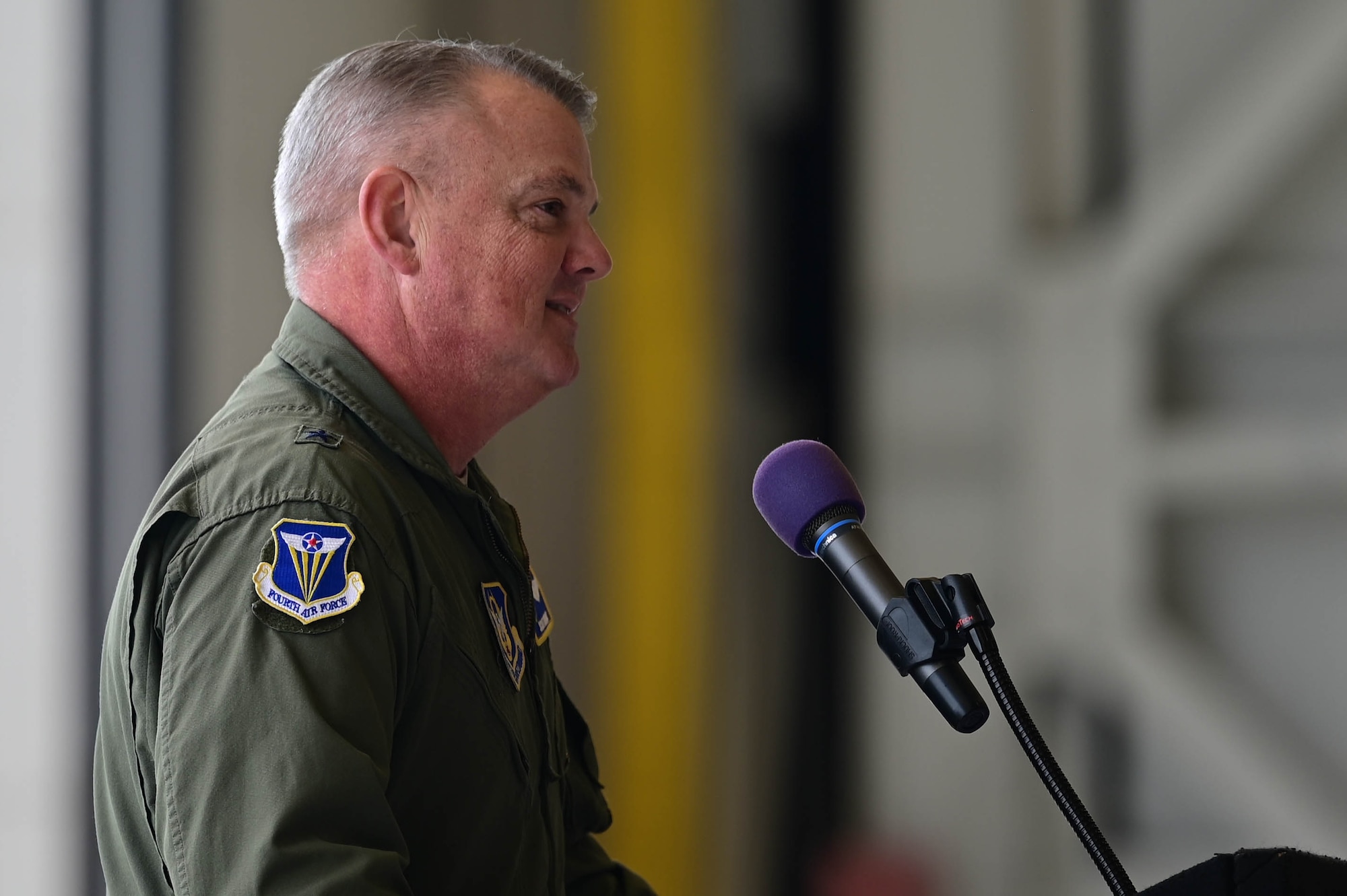 A general in a flight suit gives a speech at a podium on a stage in a hangar during a ceremony.