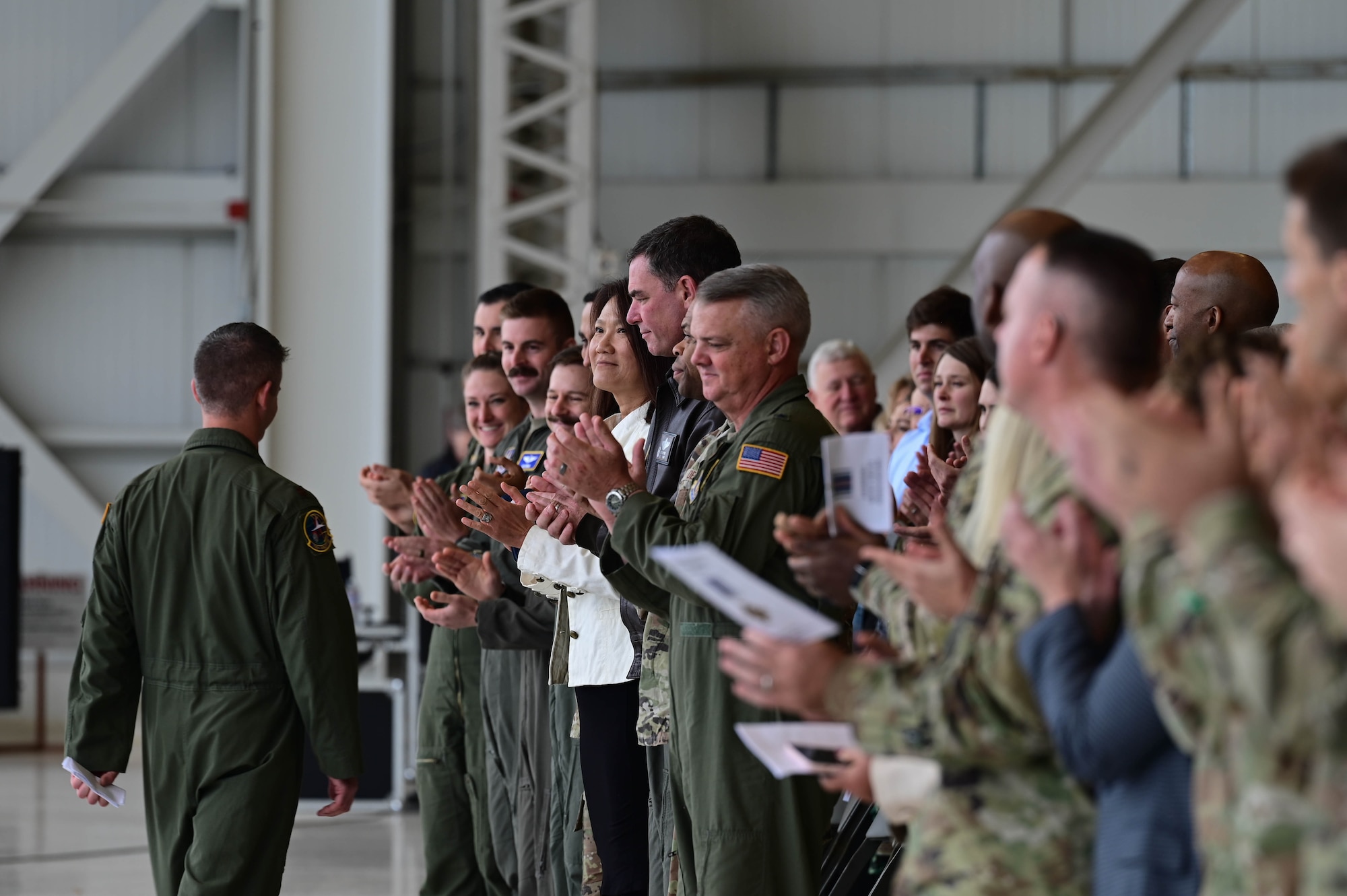 An mixed audience of Airmen, Soldiers, and civilians clap during a ceremony in a hangar.