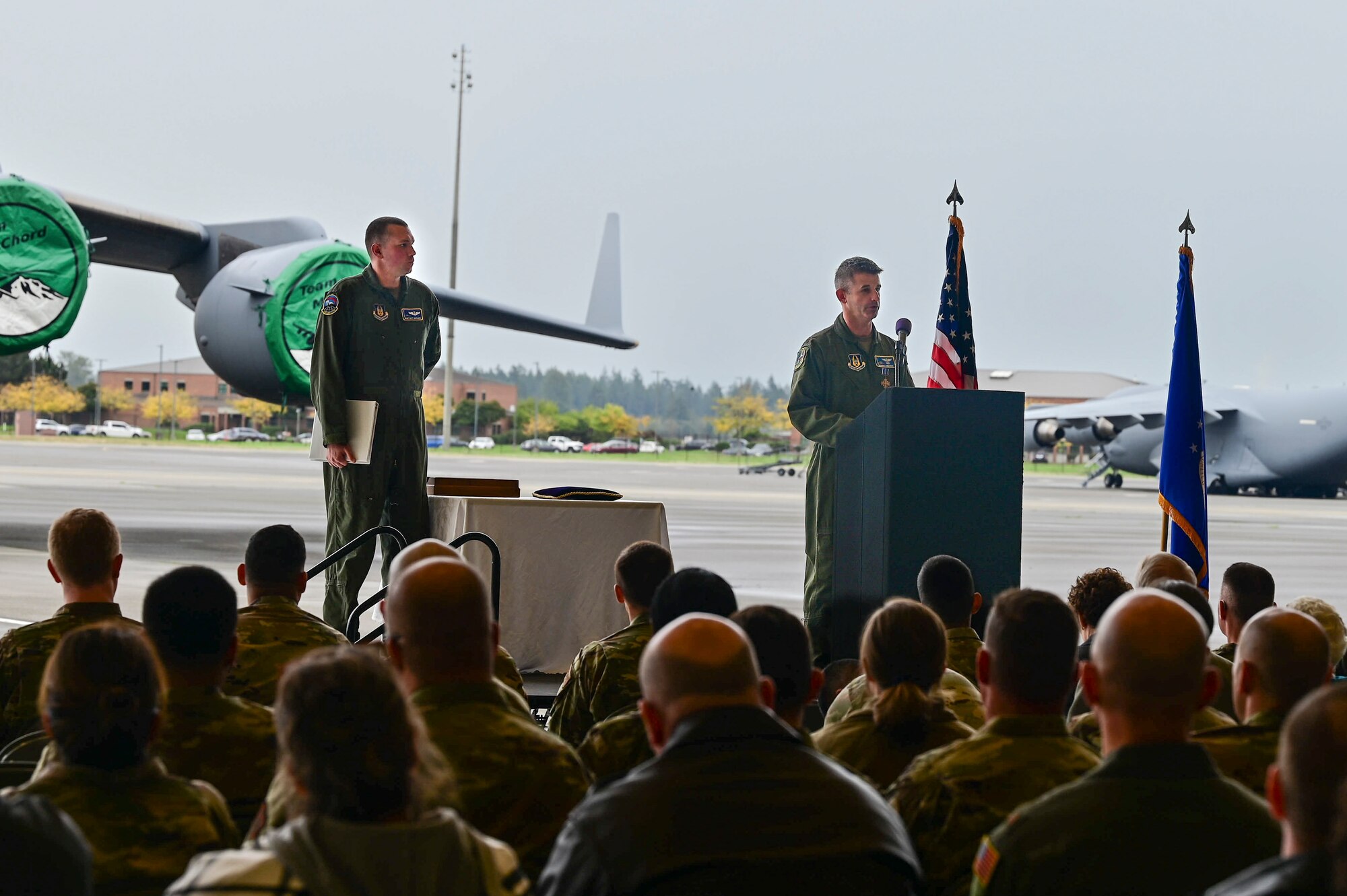 A man in a flight suit makes remarks at a podium on a stage in front of a C-17 Globemaster III to an audience.