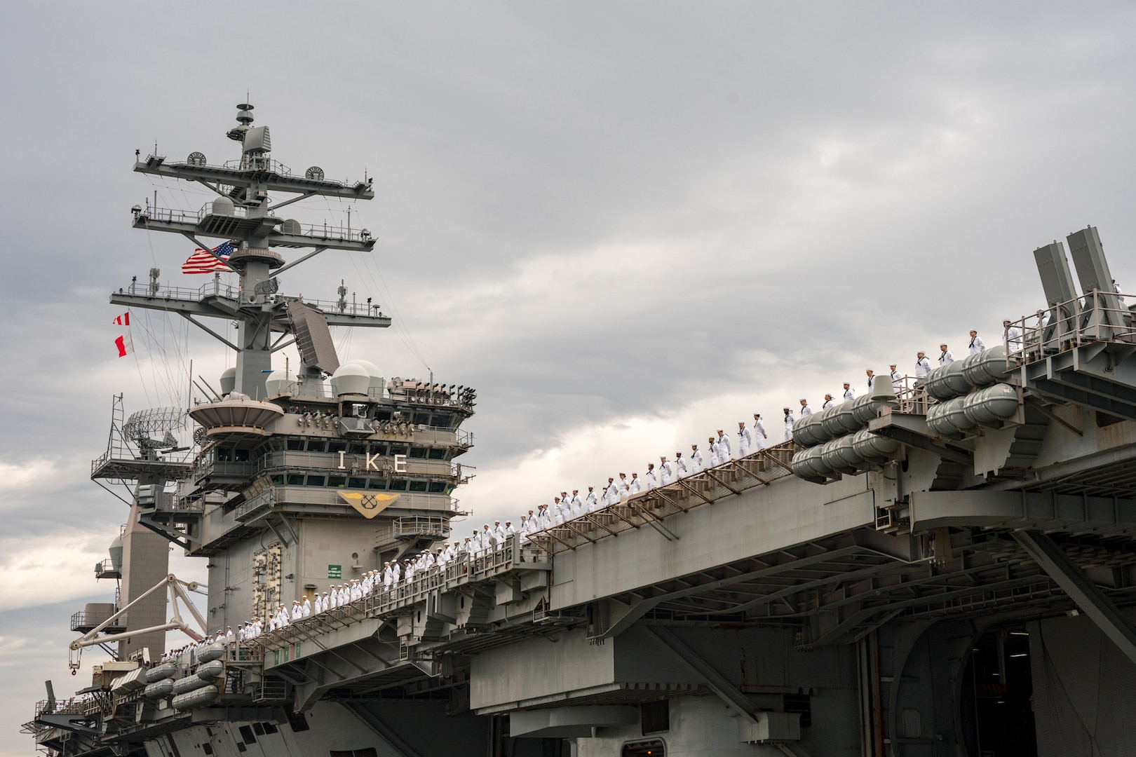 Sailors in uniform stand on the deck of an aircraft carrier.