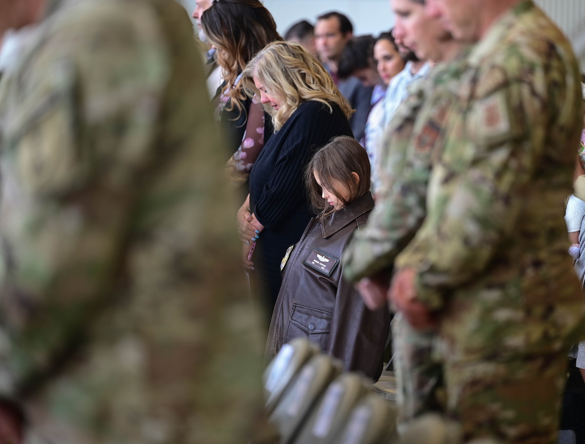 A little girl in a brown leather flight jacket bows her head during a prayer at a ceremony