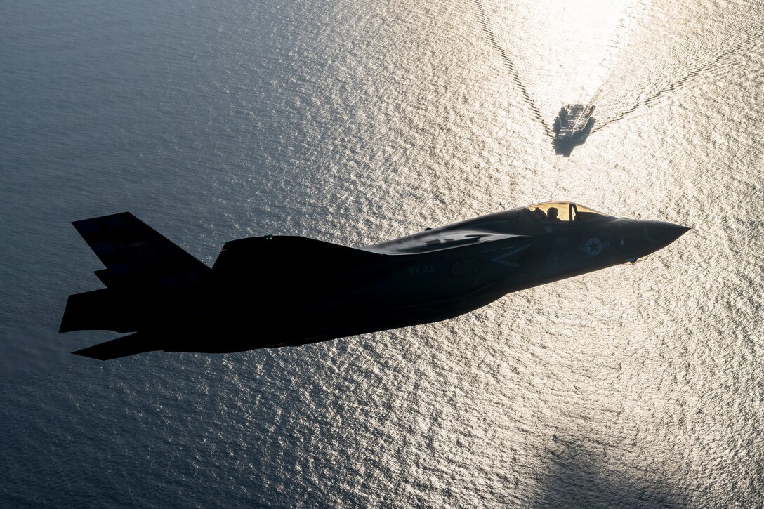 A fighter jet flies above an aircraft carrier in the ocean.