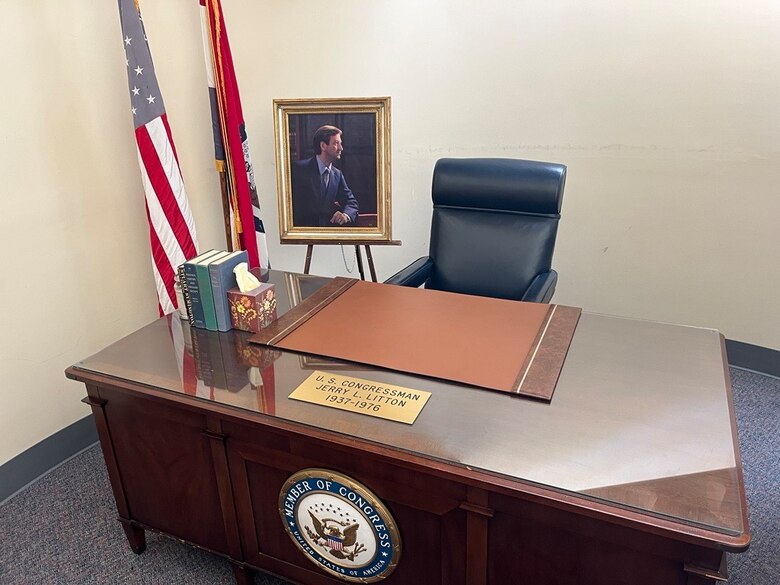 A wood desk with the U.S. Congress seal on the front, the American and Missouri State flags to the left, and a portrait in the background,