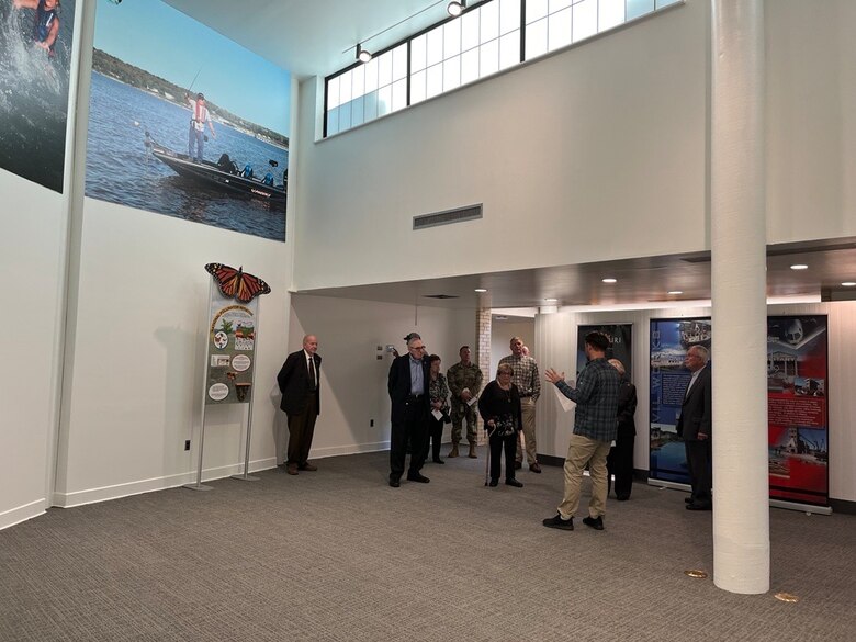 Ten people stand in an open room with murals and windows at the top of the ceiling.