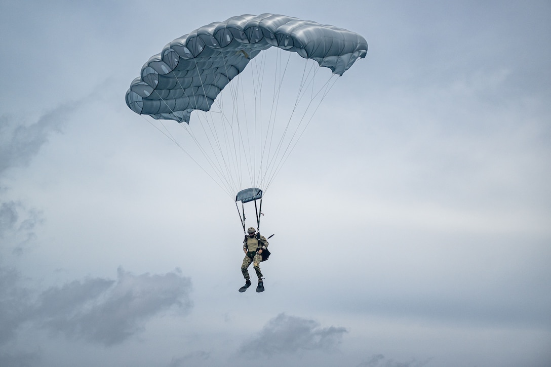 A sailor wearing a parachute descends against a cloudy sky.