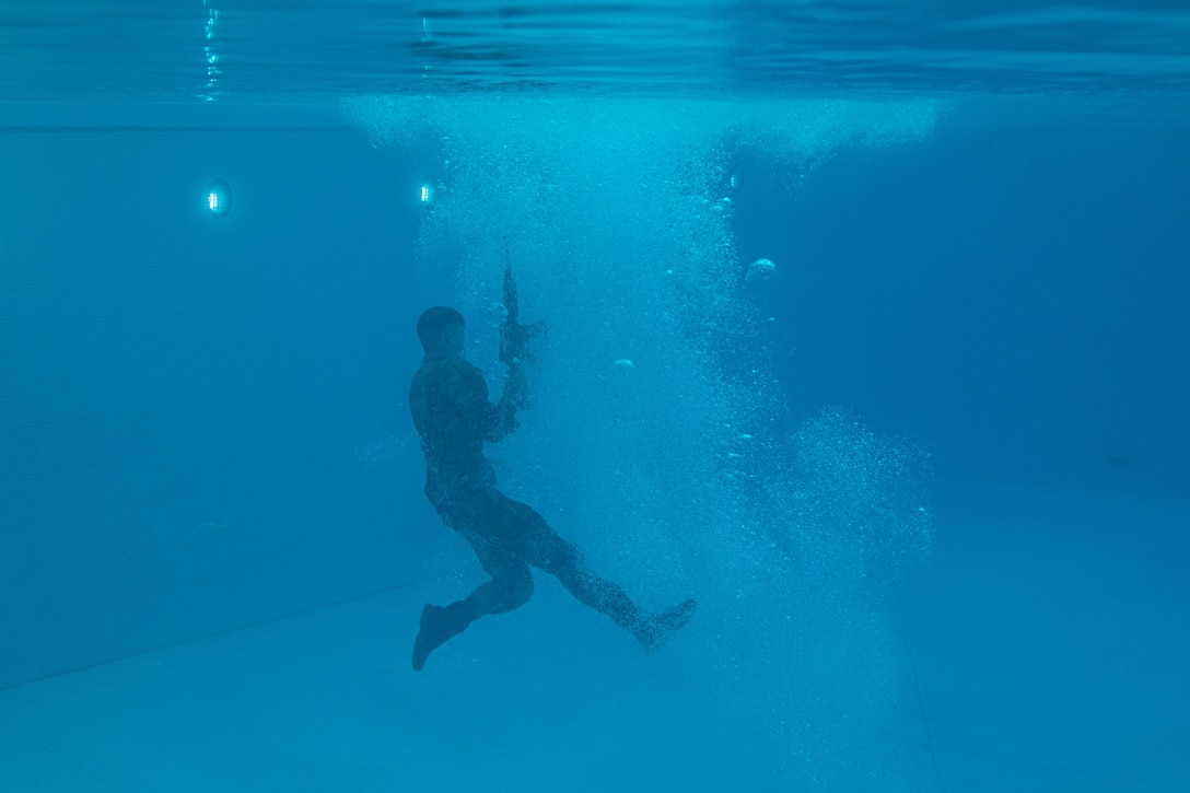 A uniformed service member holds a weapon while underwater in a pool.