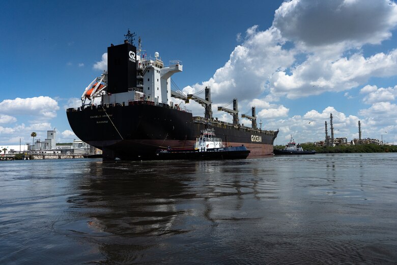 a huge cargo ship being pushed by three tugs into port