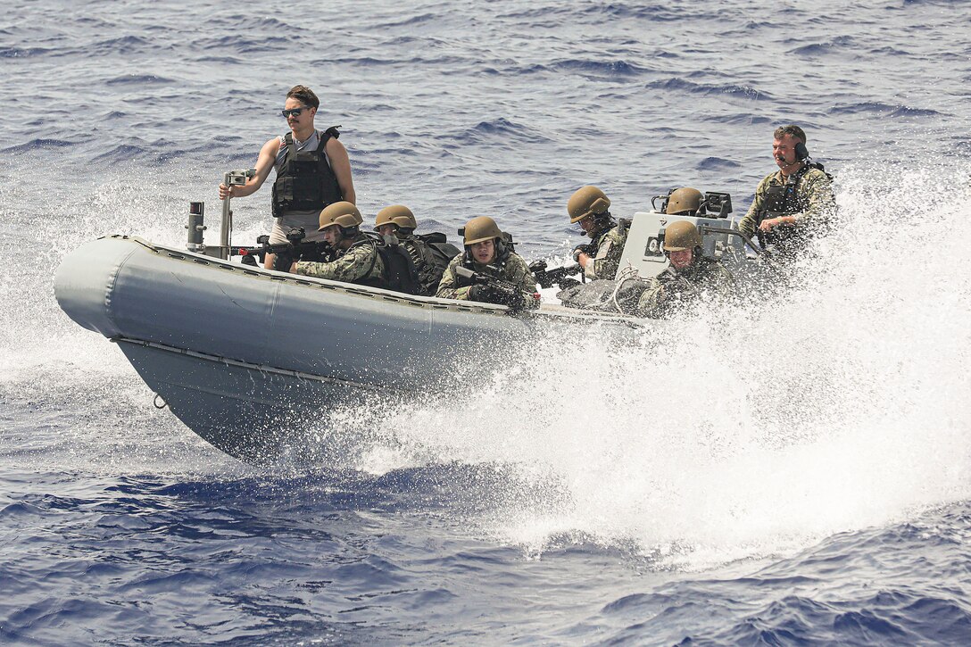 Sailors kneeling in a small inflatable boat aim weapons as water splashes around them.