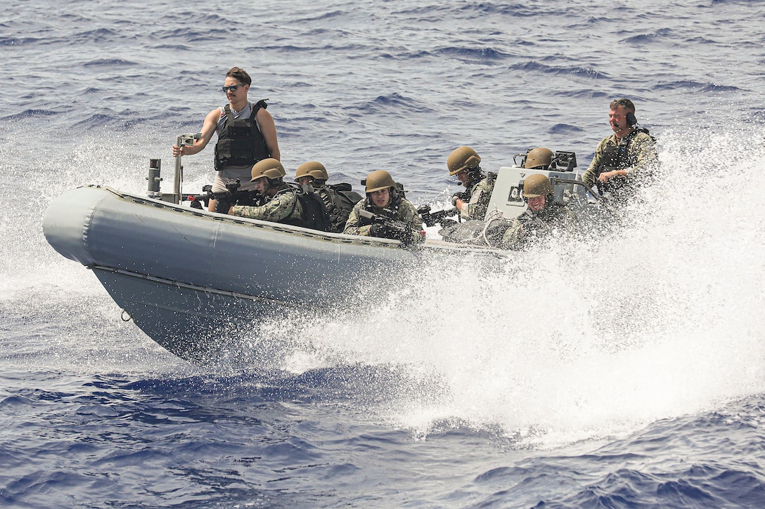 Sailors kneeling in a small inflatable boat aim weapons as water splashes around them.