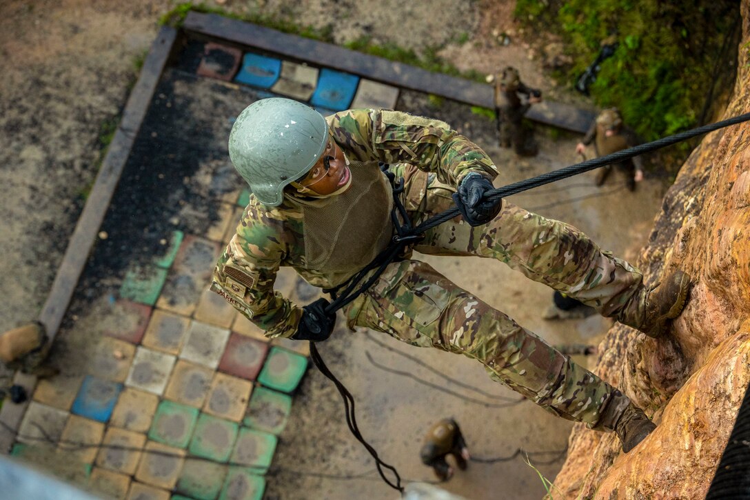 An airman rappels down a rock wall as fellow service members stand below as seen from above.