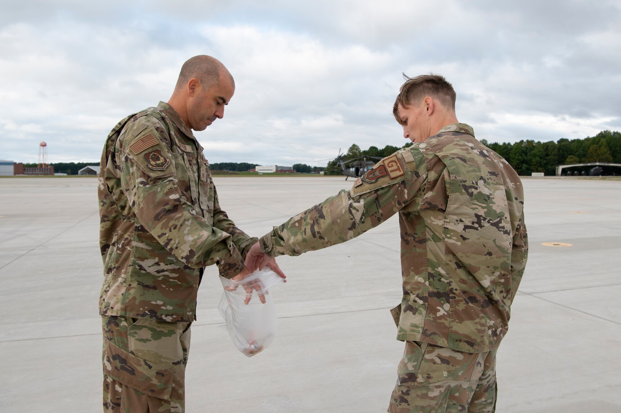 two people in military uniforms gather debris from a flightline and dispose of it into a garbage bag