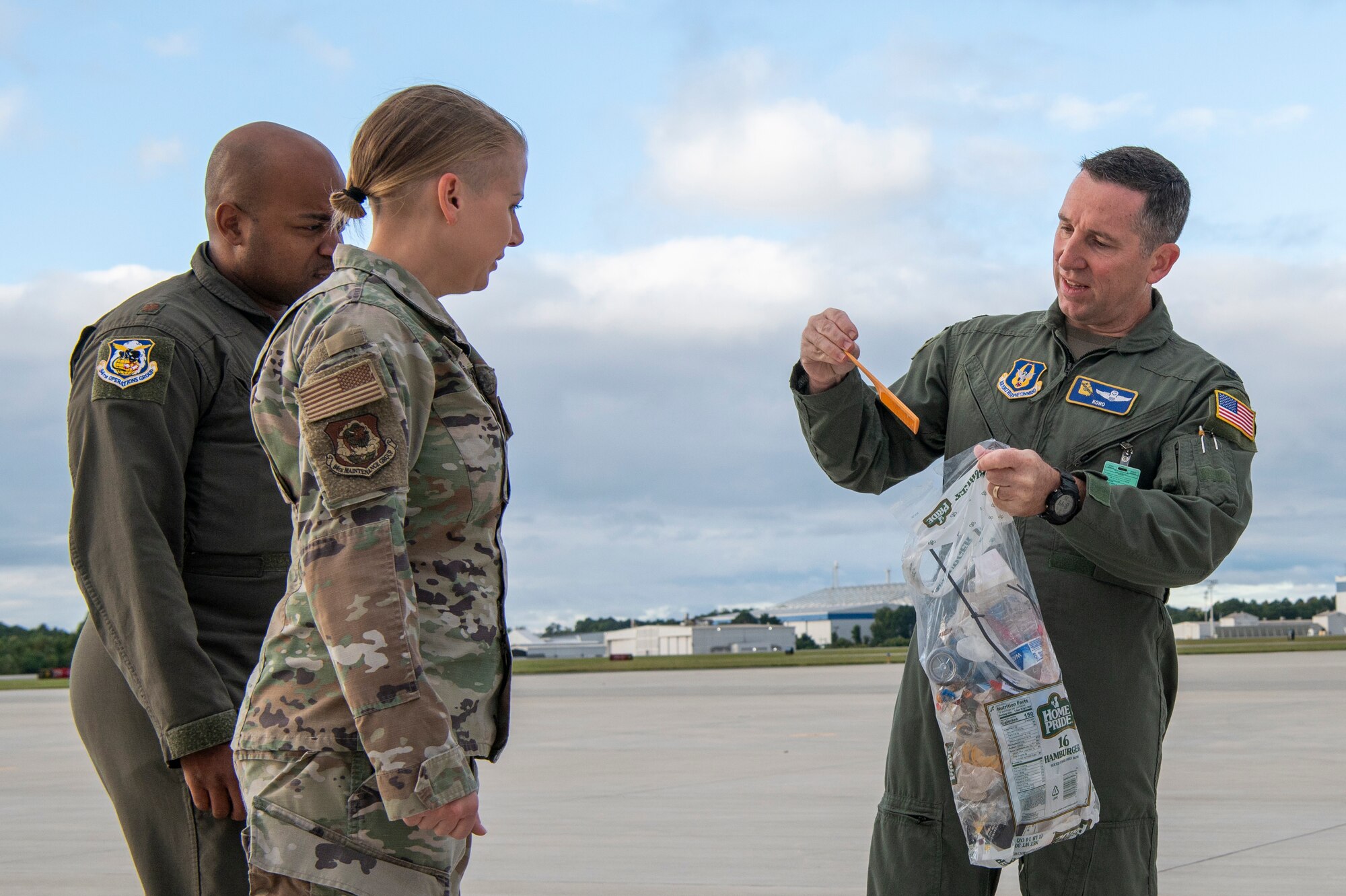 a man in a green military flight suit holds debris and a plastic bag while people in military uniforms look on