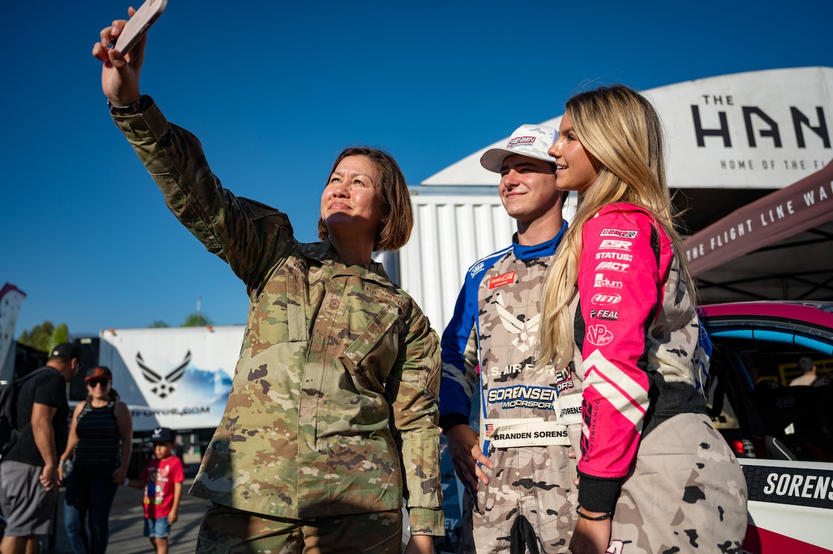 woman in uniform holds phone up into the sun to take a selfie with two race car drivers
