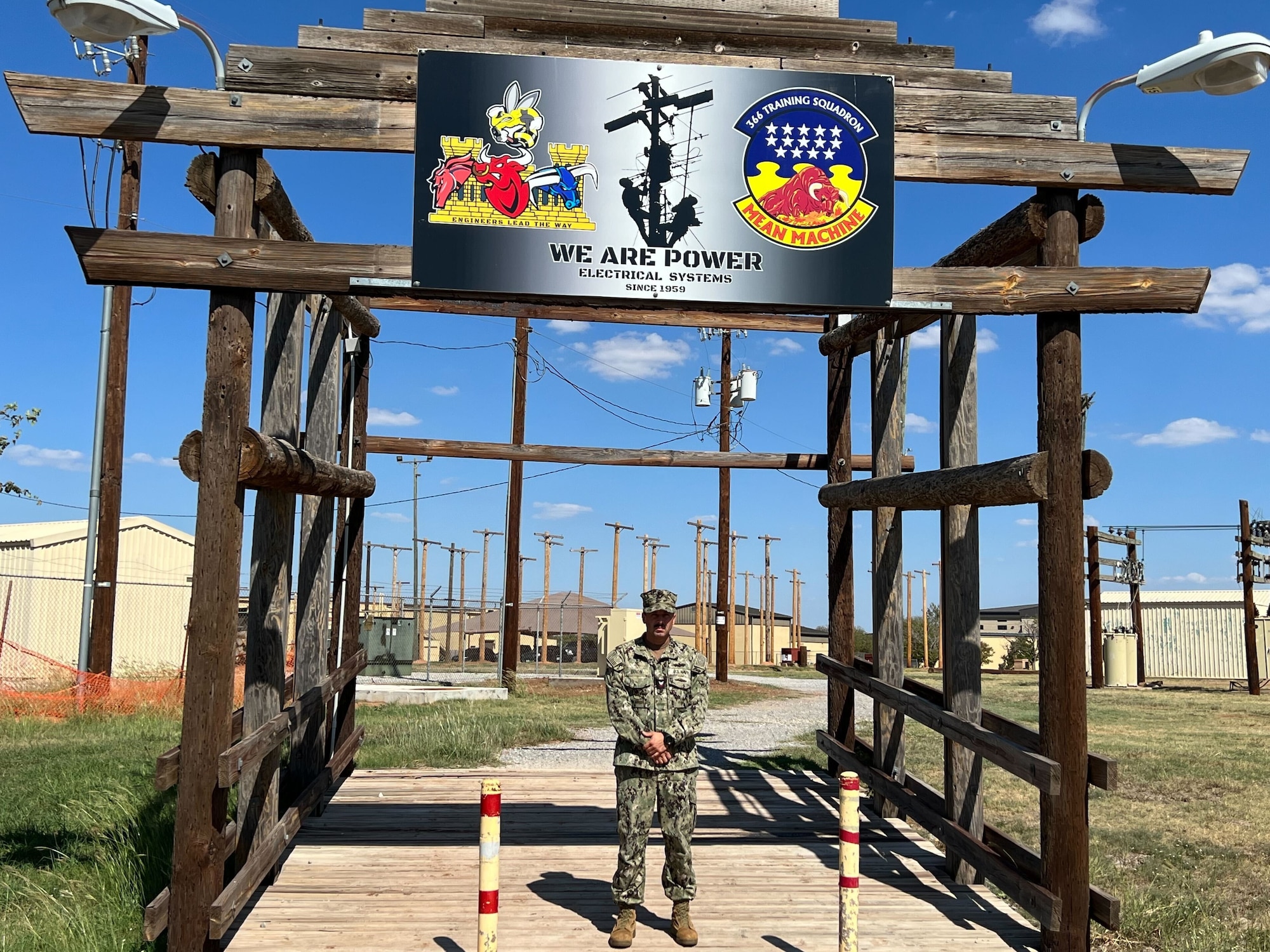 A Seabee stands under a wooden gate with utility poles in the background