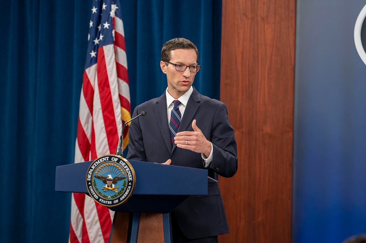 A man in a suit gestures with his hands while standing behind a lecturn.