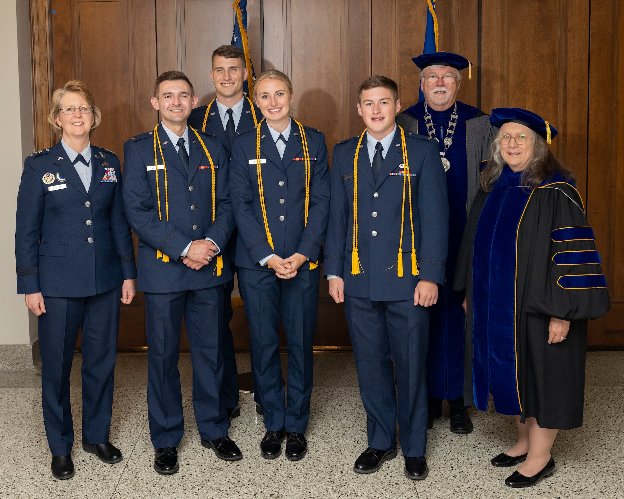 A group of three men and a woman, with a yellow cord draped from behind their necks down to their waist, pose with two people in academic robes and another woman in an Air Force uniform without a cord.