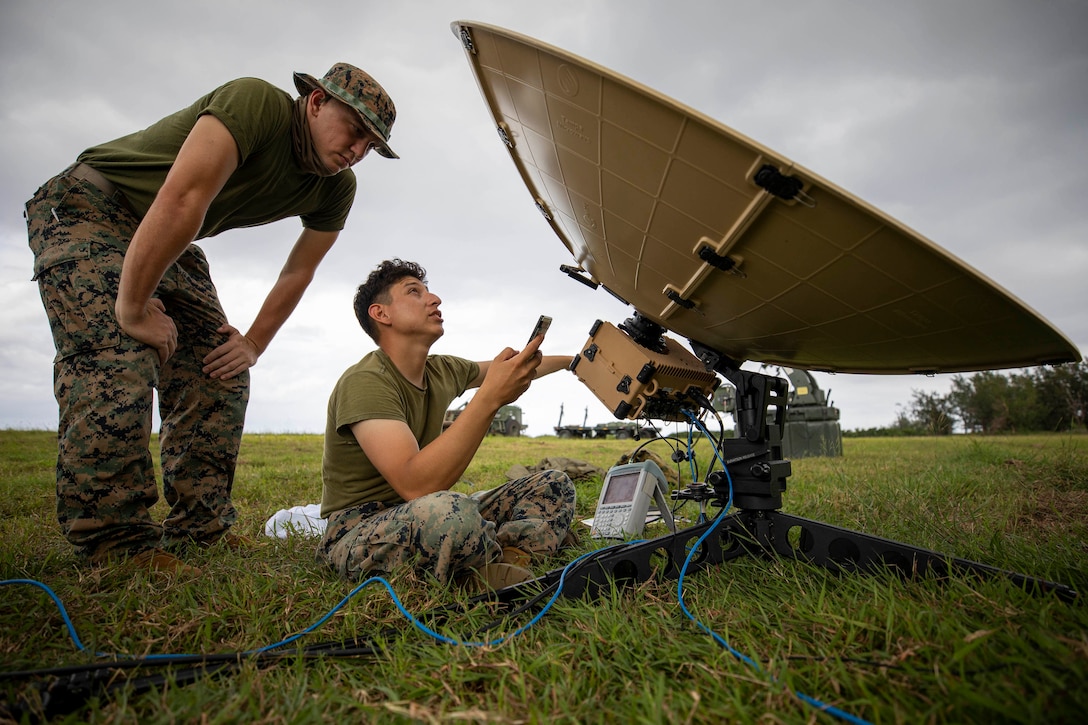 A Marine seated on grass operates a satellite dish as another watches.
