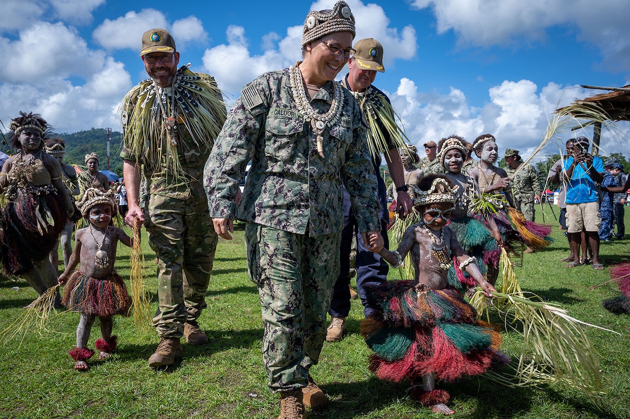 Children dressed in ceremonial attire hold hands with three uniformed service members during a community event.
