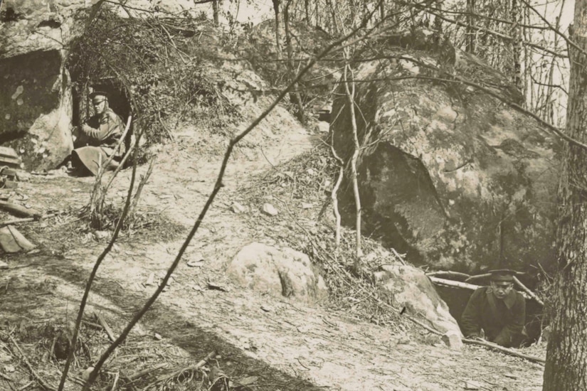 Two men hide in rock dugouts in a wooded area.