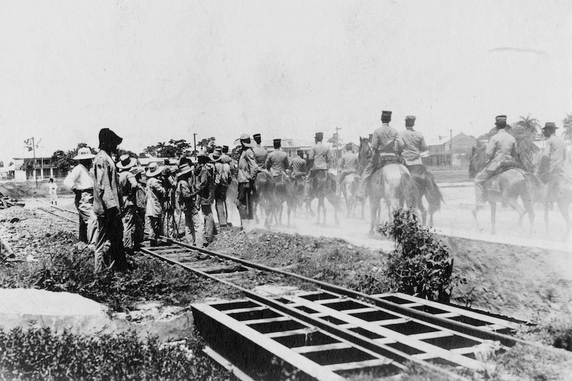 Marines ride on horses next to railroad tracks.