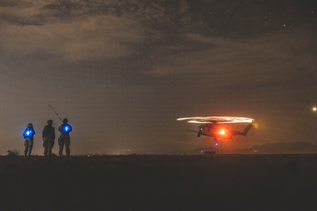 Rotator blades illuminated by white lights spin on an airborne helicopter as three Marines illuminated by blue lights stand to the left and watch under a starry sky.