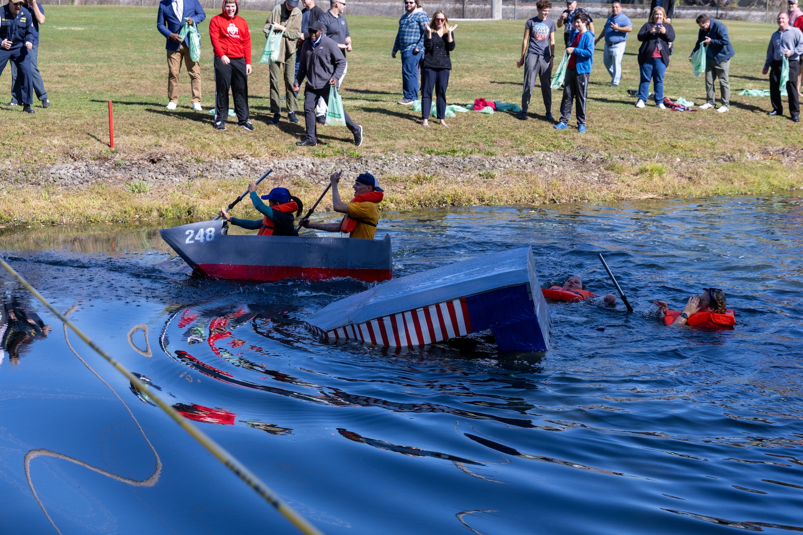 Two boats made of cardboard are racing through a pond. One boat with two rowers, a man and a woman wearing life preservers and is shaped like the hull of a ship with a Navy number the front glides past the other boat which is square and has just capsized leaving its two paddlers floating in the water with their life jackets on.