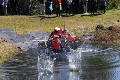 Two cardboard boats shaped like troop carriers almost and painted grey avoid water balloon munitions being thrown at them at the edge of the pond that served as the starting line for a boat race. The four Navy men in a hodge podge of various Navy type dress are furiously paddling to get away from the fray.