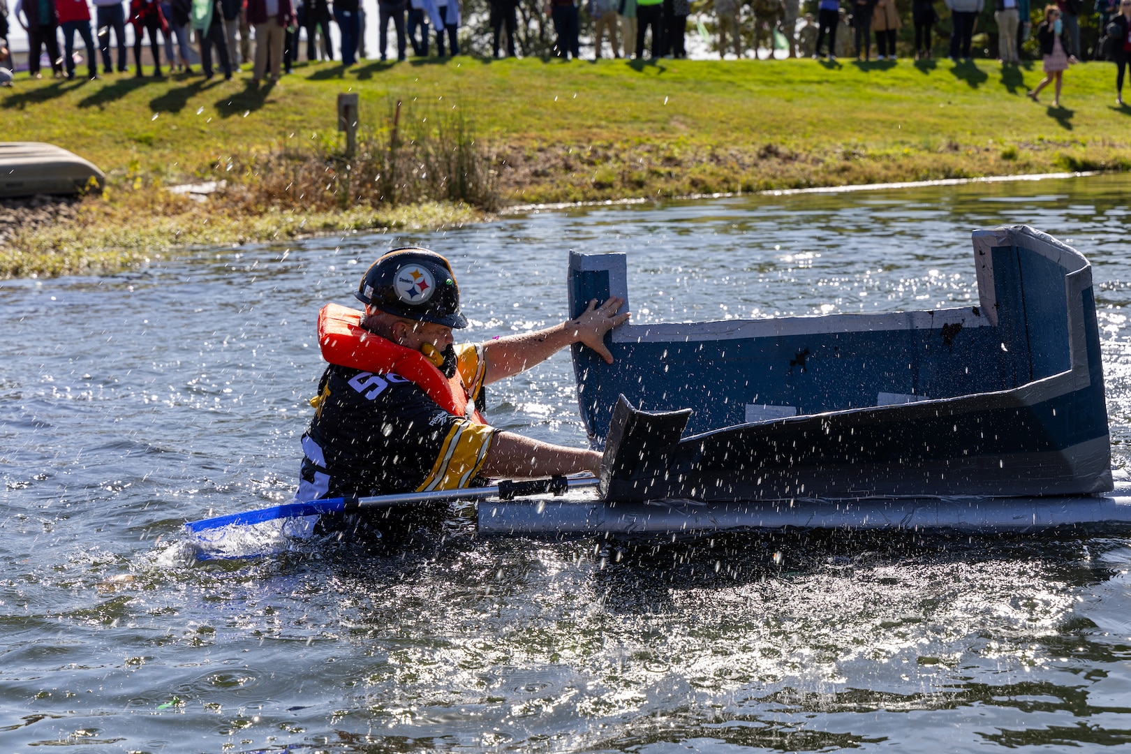 A man in a Steelers outfit tries to get aboard his cardboard boat in a pond after it capsized. The boat is square with an open back and is gray.