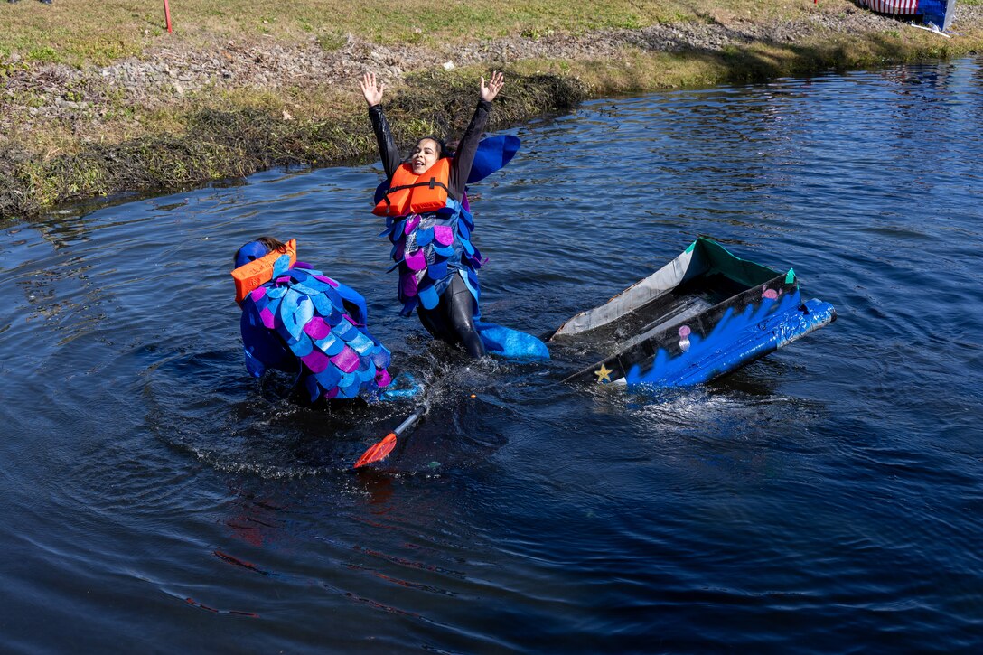Two women in blue, green and silver fish suits fall out of their half sinking cardboard boat which is boxy and blue with low sides in a pond.