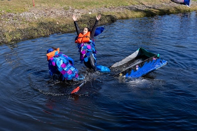 Two women in blue, green and silver fish suits fall out of their half sinking cardboard boat which is boxy and blue with low sides in a pond.