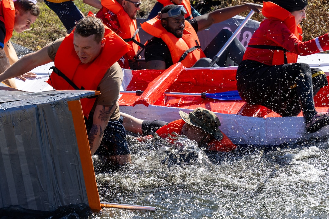 Men and women with various costumes and life preservers on scramble to get into their cardboard boats at the beginning of a race at the edge of a pond. One man is dark skinned and one man wears a green boonie hat. A woman in a red and white Olive Oyl dress is almost out of the shot.