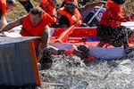Men and women with various costumes and life preservers on scramble to get into their cardboard boats at the beginning of a race at the edge of a pond. One man is dark skinned and one man wears a green boonie hat. A woman in a red and white Olive Oyl dress is almost out of the shot.