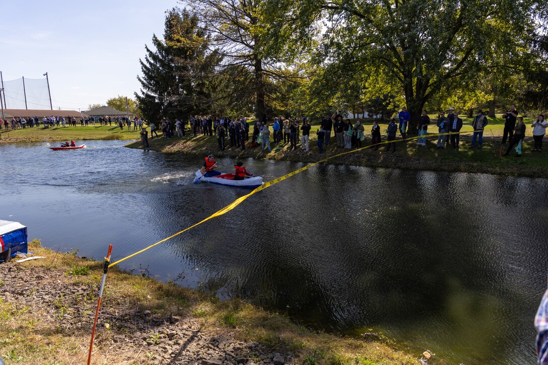 A red, white and blue canoe-shaped cardboard boat glides through the water in a pond with many people looking on.