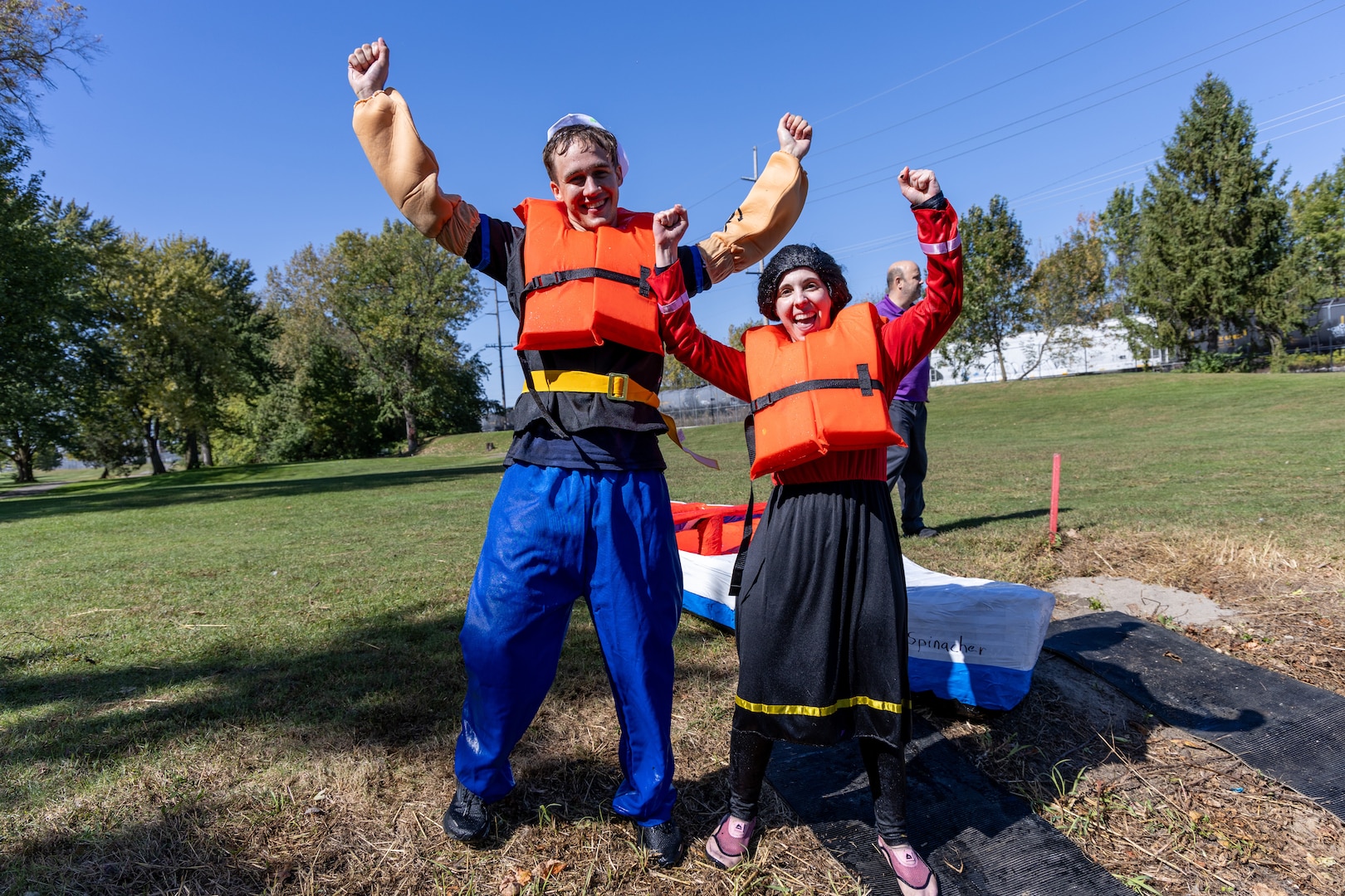 A man and a woman dressed in Popeye the Sailor Man and Olive Oyl costumes complete with the white sailor hat and black hair wig and a red and black dress have their hands up in the air celebratory style. A large canoe-shaped cardboard boat sits behind them. They are outside.