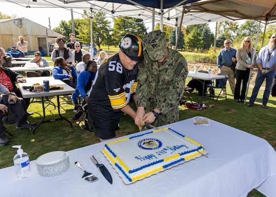 A man in a Steelers helmet and jersey cuts a cake with a sword along with a younger man in a camouflaged military uniform. They are the only ones at a table with a white tablecloth under a tent outside.