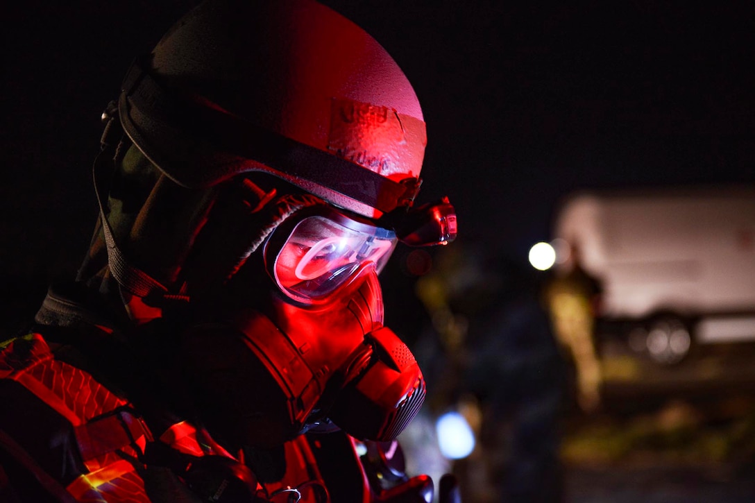 A service member in protective gear looks forward and is illuminated by a red light.