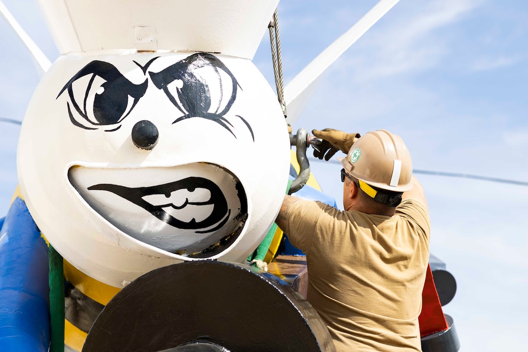 A sailor attaches crane rigging to a large bee statue with a smirking, human facial expression.