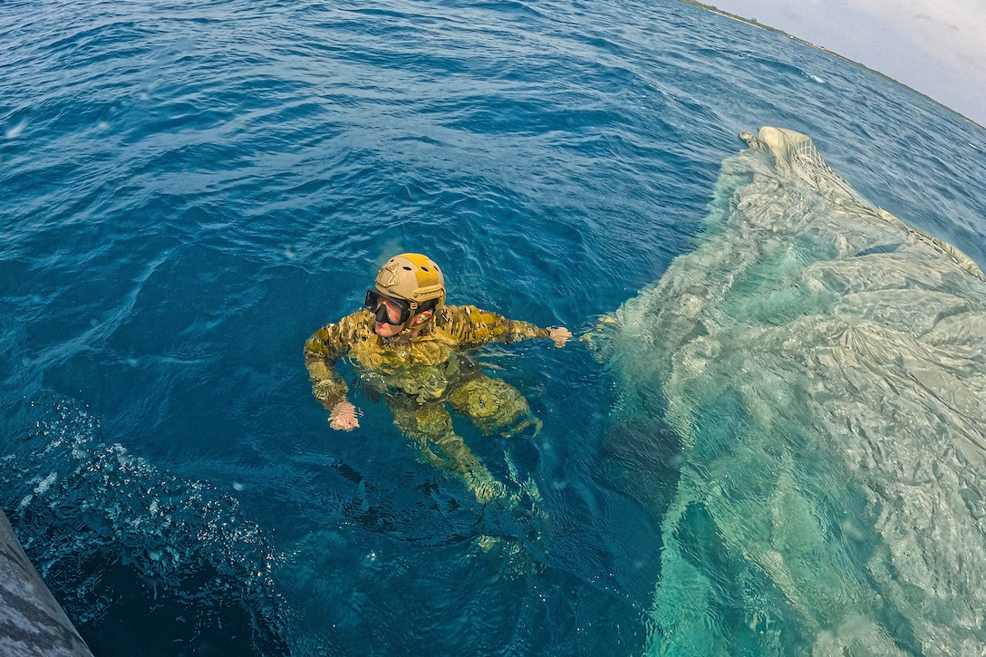 A sailor wearing goggles treads water attached to a parachute submerged in water to the right.