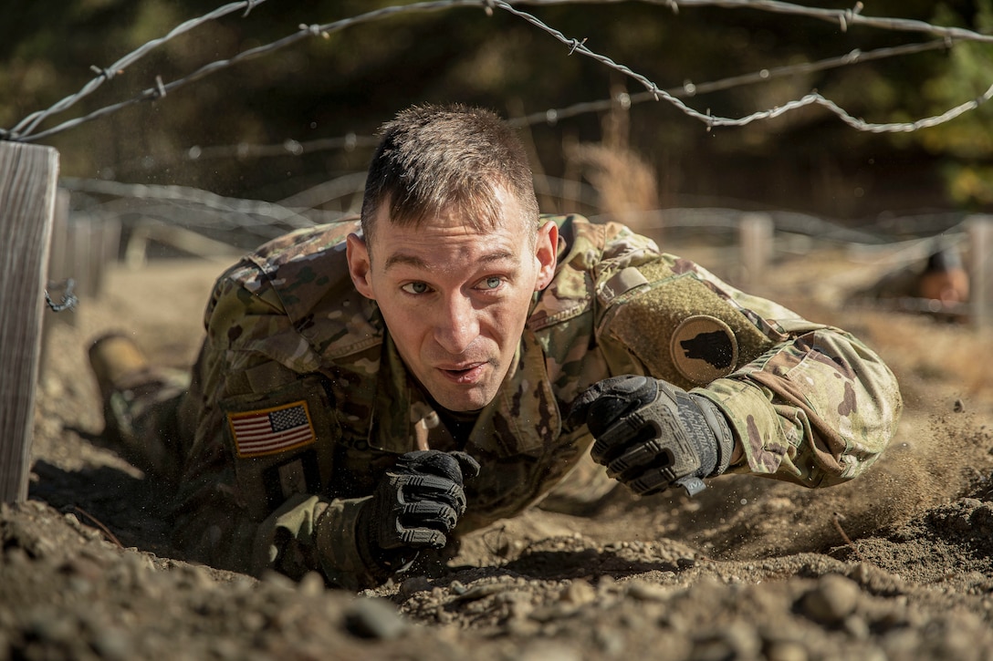 A soldier crawls in the dirt under barbed wire.
