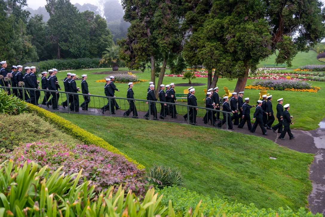 Dozens of sailors walk down a ramp surrounded by manicured lawns with colorful flowers and trees.