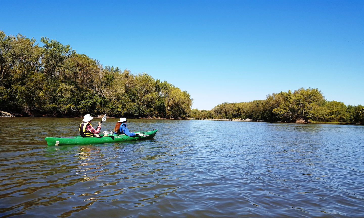 Two people are seated in a green kayak on the water with trees in the background.