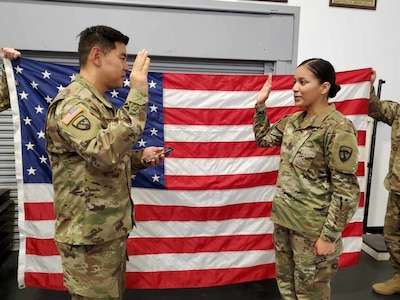 A man and woman, wearing Army uniforms, stand in front of the American Flag with their right hands up.