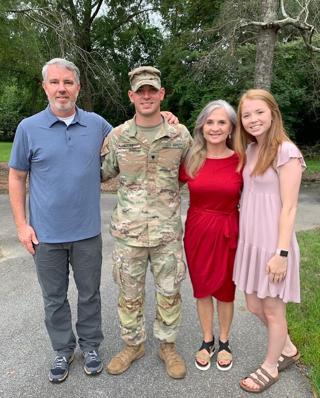 Man in a blue polo and jeans, a man in an Army uniform, a woman in a red dress, and a woman in a pink dress pose together in front of trees