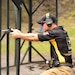 Man in Army polo, with ear protectors on, takes a knee while aiming a gun during a marksmanship exercise