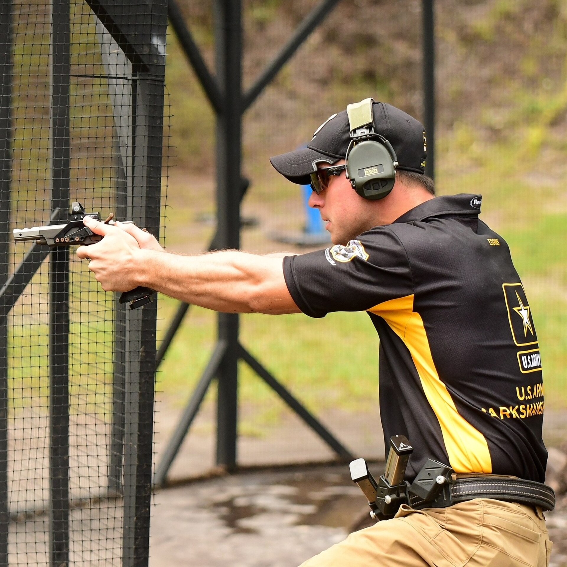 Man in Army polo, with ear protectors on, takes a knee while aiming a gun during a marksmanship exercise
